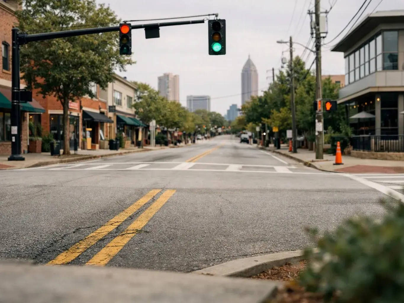 Empty downtown street stretches under green traffic lights, with storefronts, trees, and distant high-rise buildings